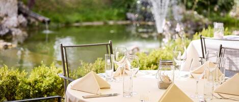 An outdoor dining area with round tables set for two, each with a white tablecloth, glassware, and cutlery.