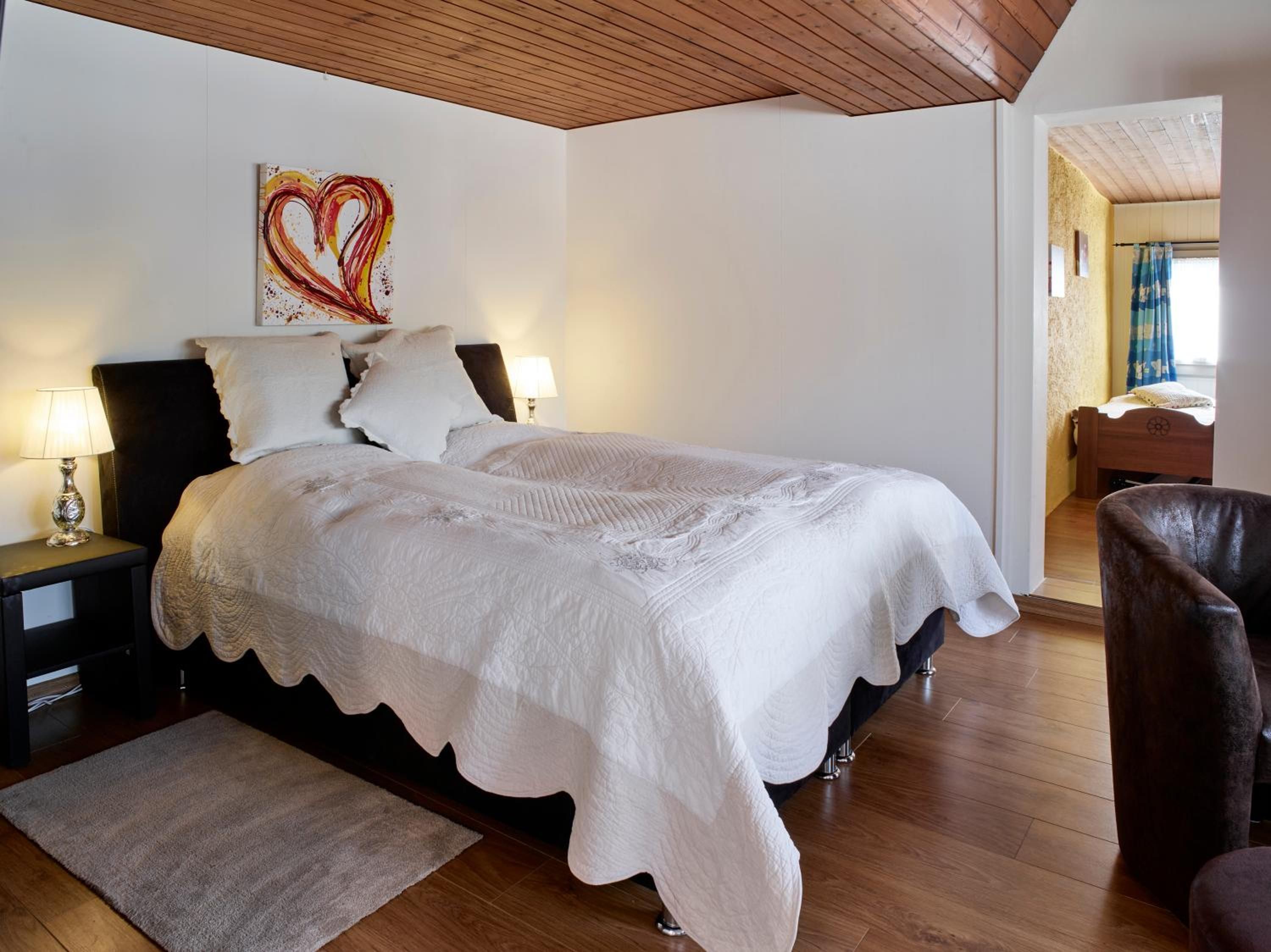 Ein Schlafzimmer mit einem Bett, Nachttischen, einer Lampe, einem herzförmigen Gemälde und einer Holzbalkendecke.