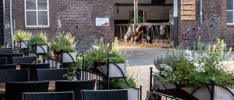 An outdoor seating area with wicker furniture and potted plants, a brick building with a sign, and a cow feeding area visible through an open doorway.