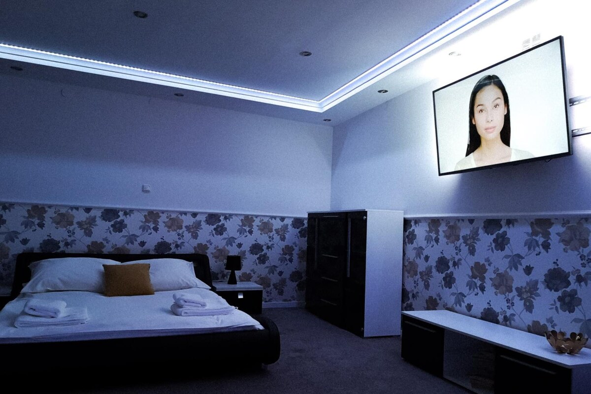 Un dormitorio con una cama, televisor montado en la pared, cómoda y un banco.