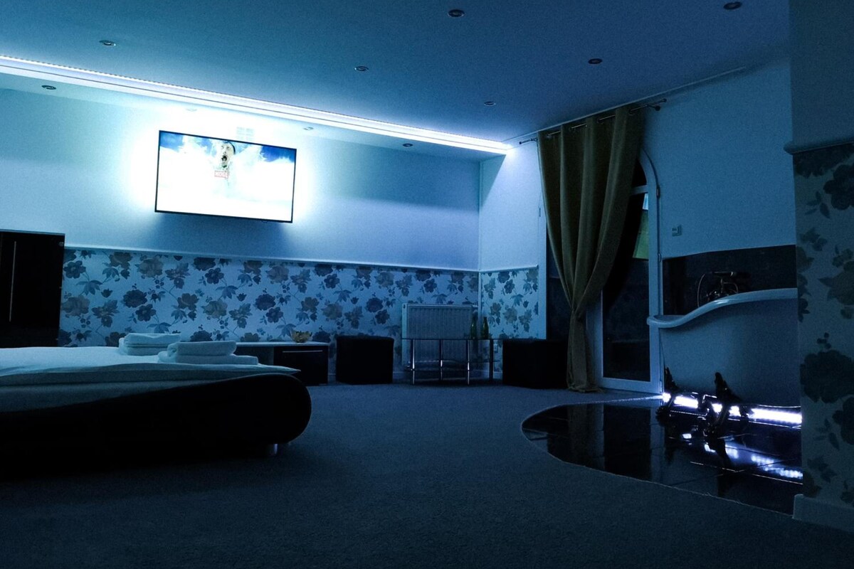A hotel room with a bed, a flat-screen TV mounted on the wall, a mirror, and a window with curtains.