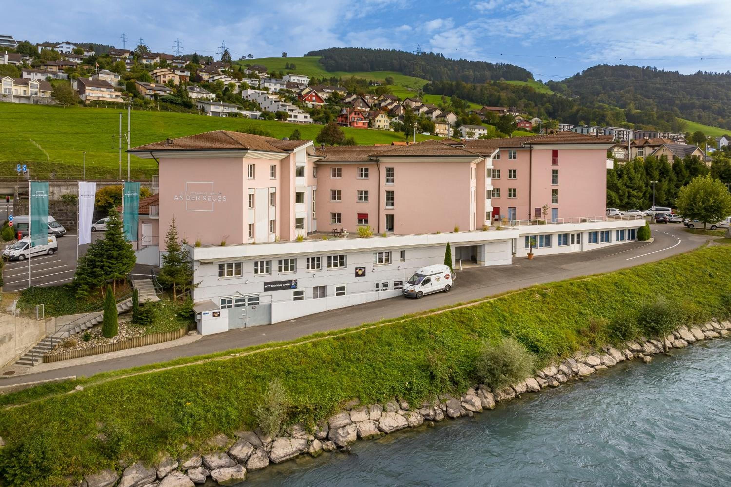 Un hotel con una fachada moderna, ubicado junto a un río, y vista a un pueblo montañoso al fondo.
