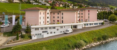 Un hotel con una fachada moderna, ubicado junto a un río, y vista a un pueblo montañoso al fondo.