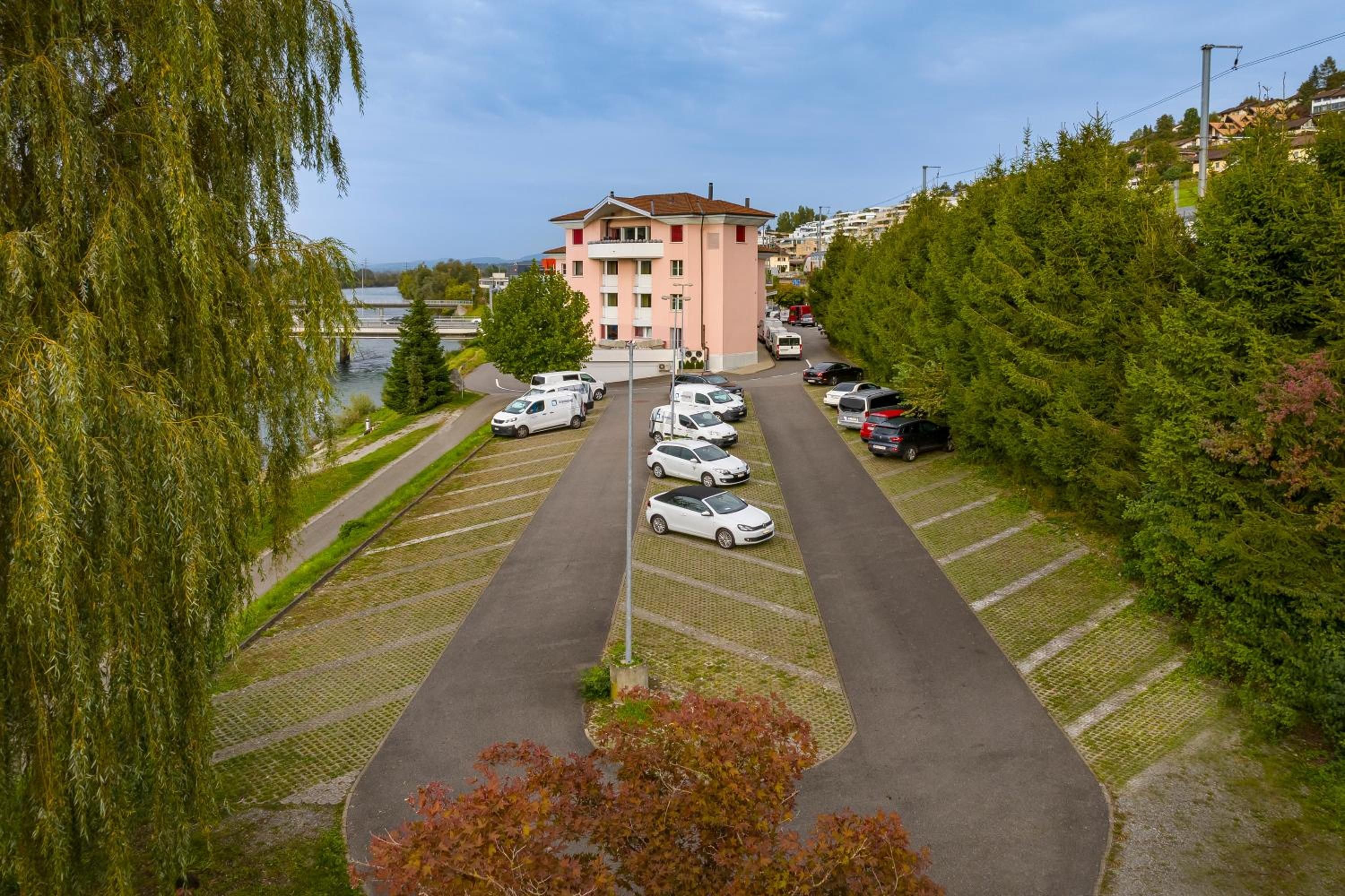 A parking lot with multiple cars, a building with a red roof, and a river in the background.