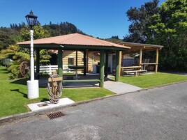 A covered picnic area with a bench and a table, surrounded by a grassy lawn and trees.