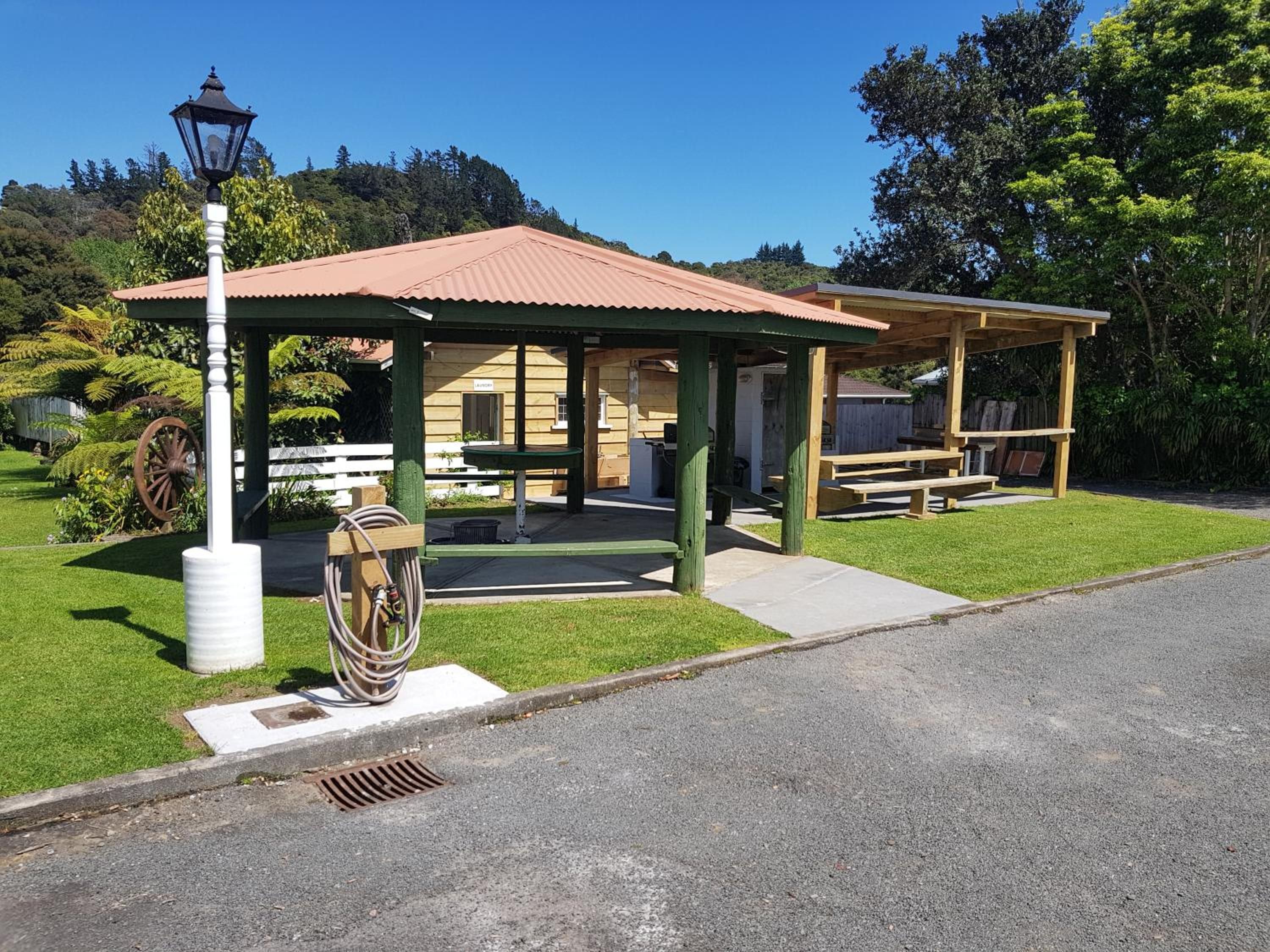 A covered picnic area with a bench and a table, surrounded by a grassy lawn and trees.