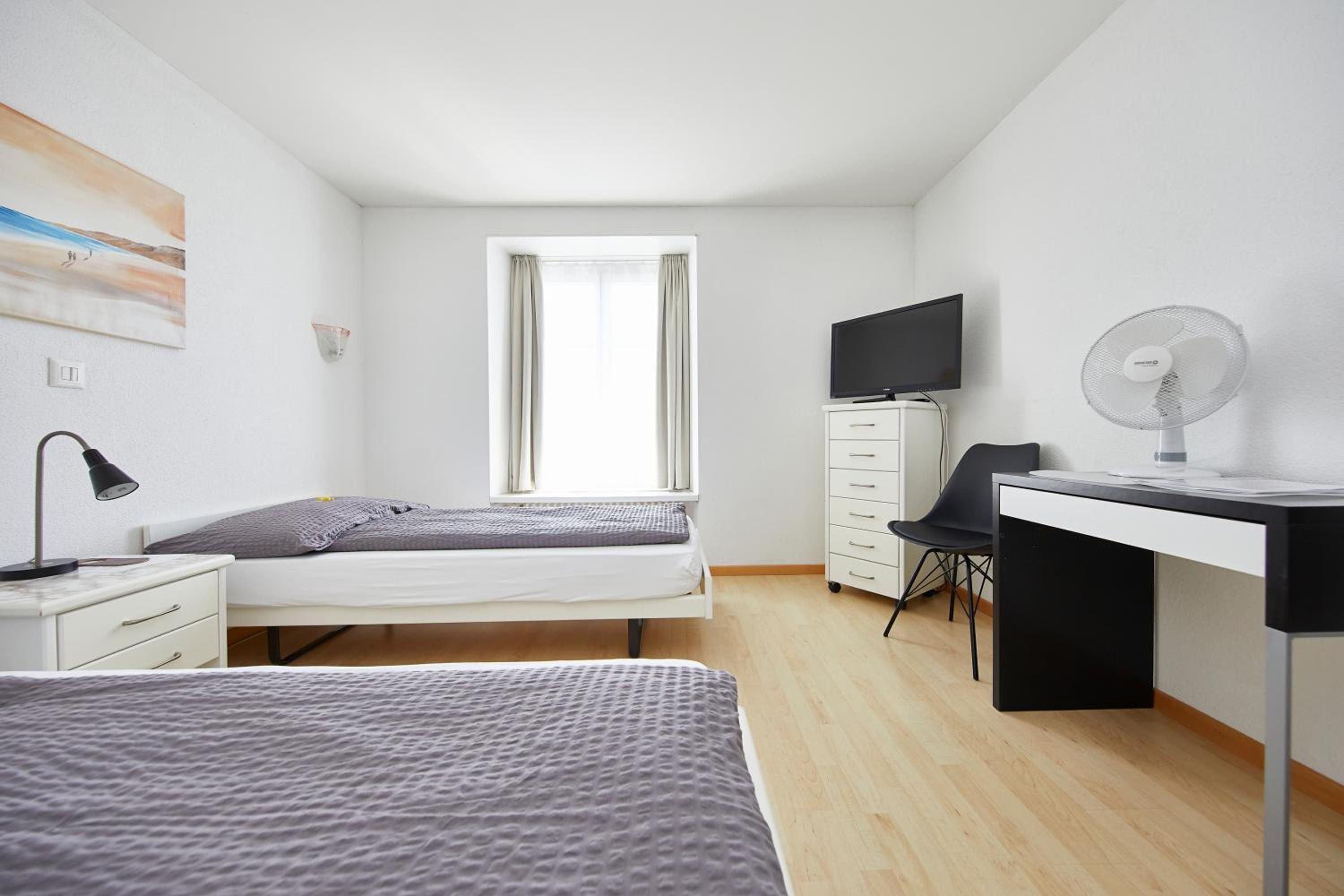 A bedroom with a bed, a desk with a chair, a TV, a lamp, and a window.
