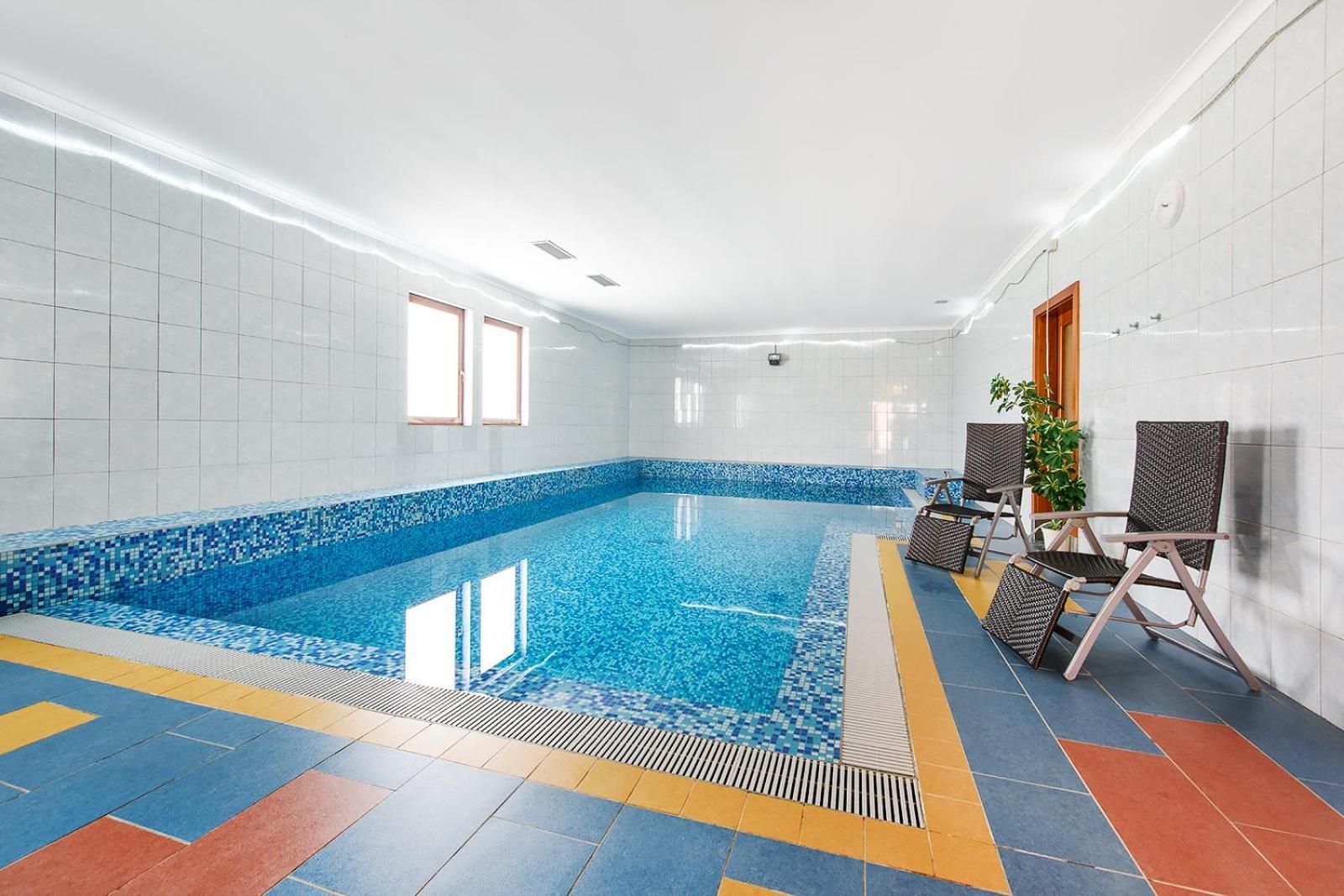Ein Hallenbad mit gefliesten Wänden, einer blauen Mosaikbordüre und einem Boden mit gelb-rotem Muster. Zwei Fenster und ein Blumentopf befinden sich in der Nähe des Pools.