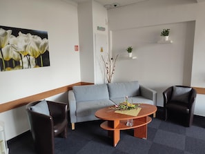 Ein Empfangsbereich mit grauem Sofa, zwei Sesseln, einem Holztisch mit Blumenarrangement, einer Wandregal für Pflanzen und einem Brandschutztürschild.