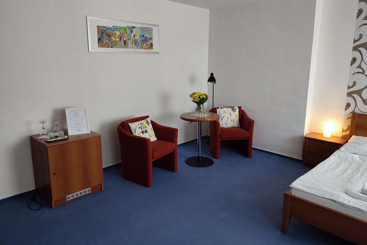 Habitación de hotel con una cama, dos sillones rojos, una mesita redonda con una lámpara, un jarrón con flores y una obra enmarcada en la pared.