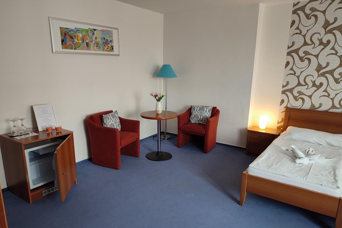 Ein Hotelzimmer mit einem Bett, zwei Sesseln, einem kleinen Tisch mit Blumenvase, einer Lampe, einem Schreibtisch mit Speisekarte und einem Wandbild.