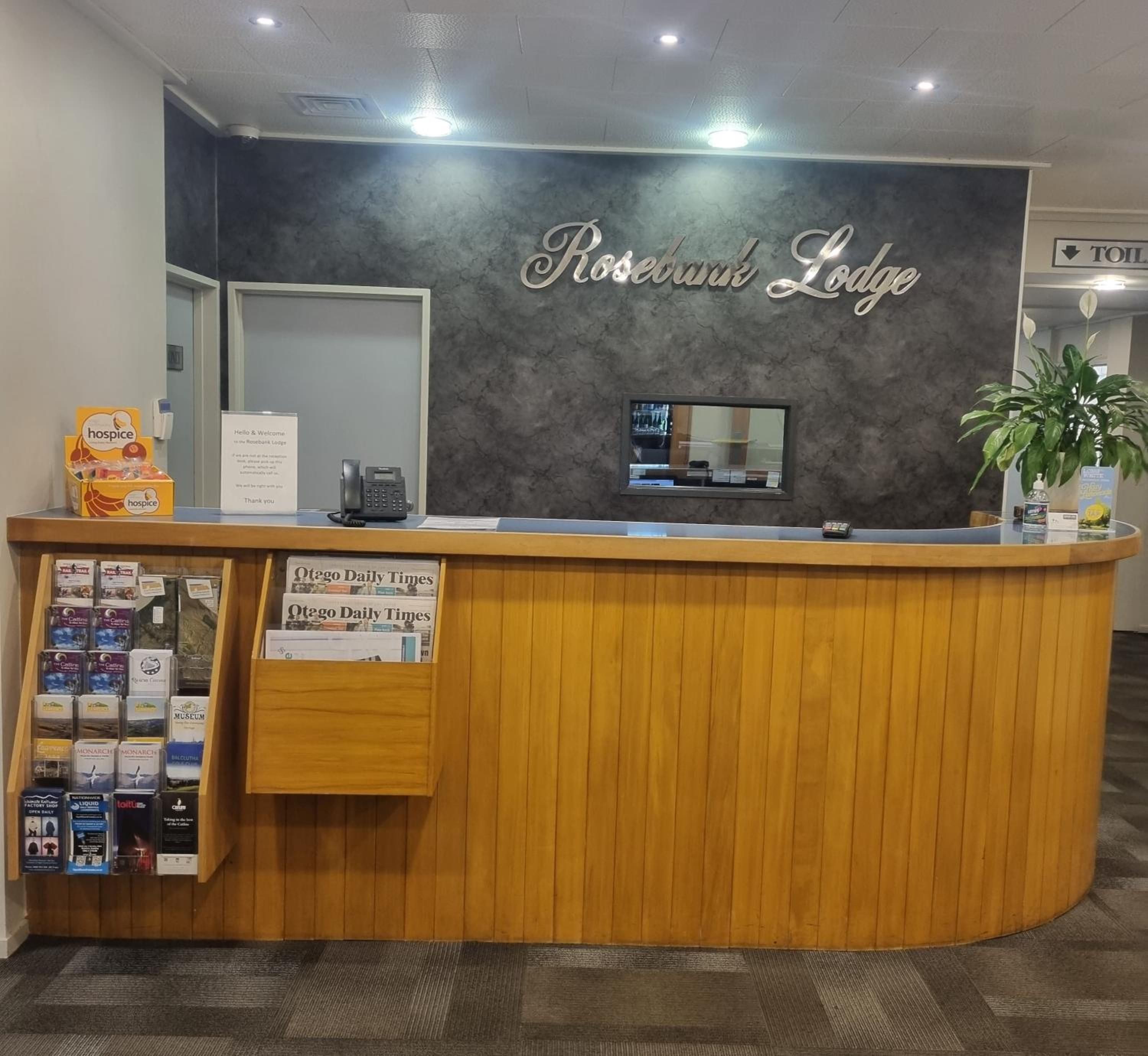 A reception desk with a sign reading 'Rosebank Lodge' and a display of local newspapers.