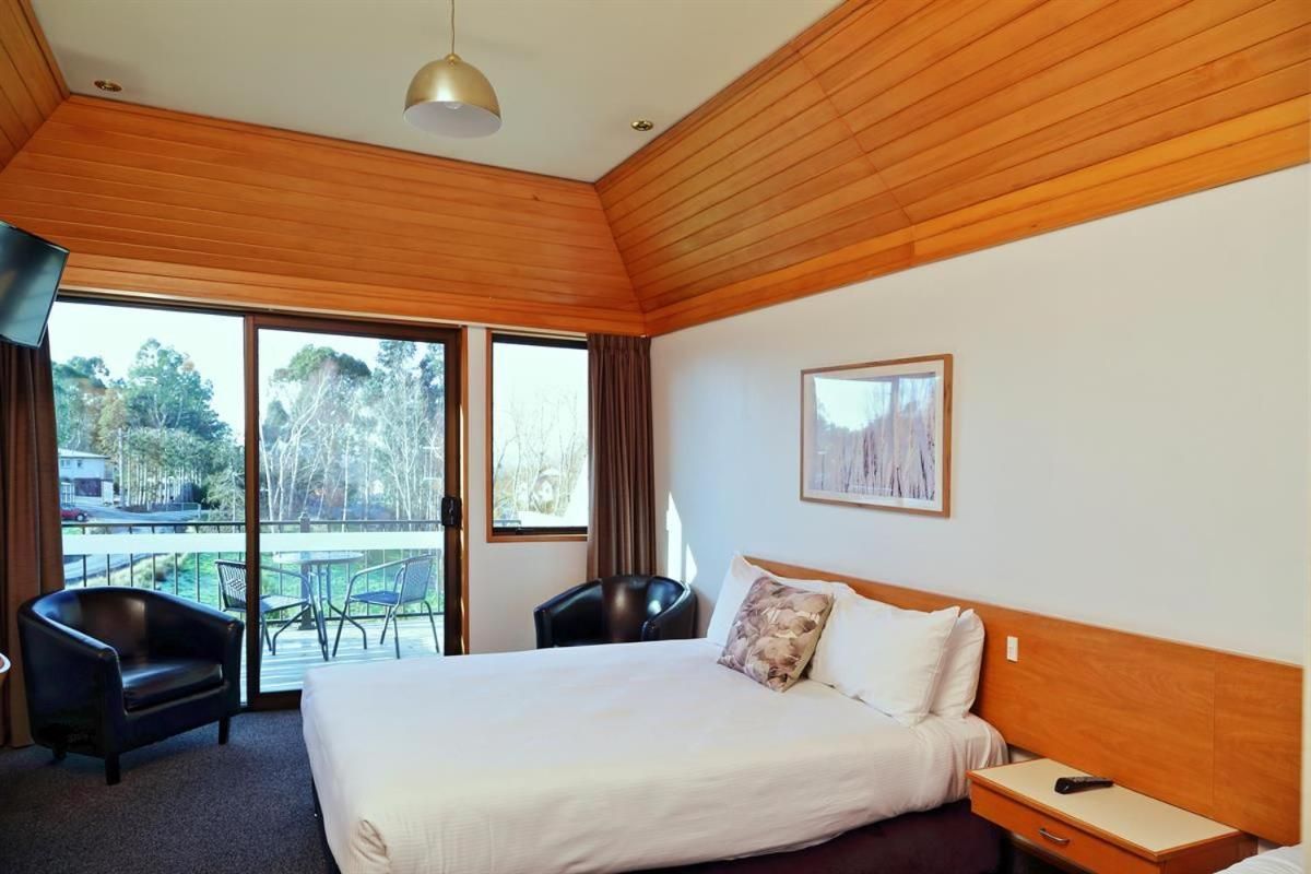 A hotel room with a large bed, two chairs, a nightstand, and a window with a view of trees and a balcony.