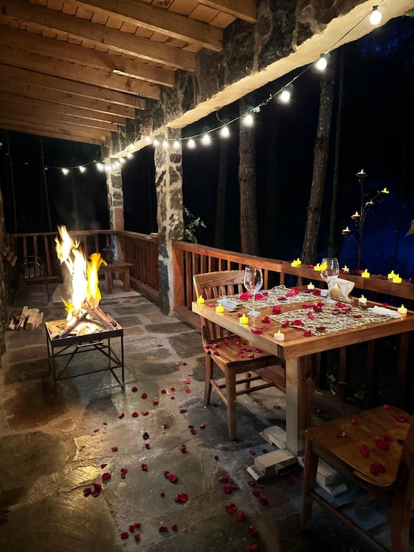An outdoor dining area with a fire pit, string lights, and scattered rose petals on the stone floor.