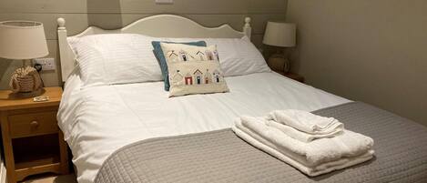 A neatly made bed with a quilted bedspread, a decorative pillow, and folded towels on a wooden nightstand.