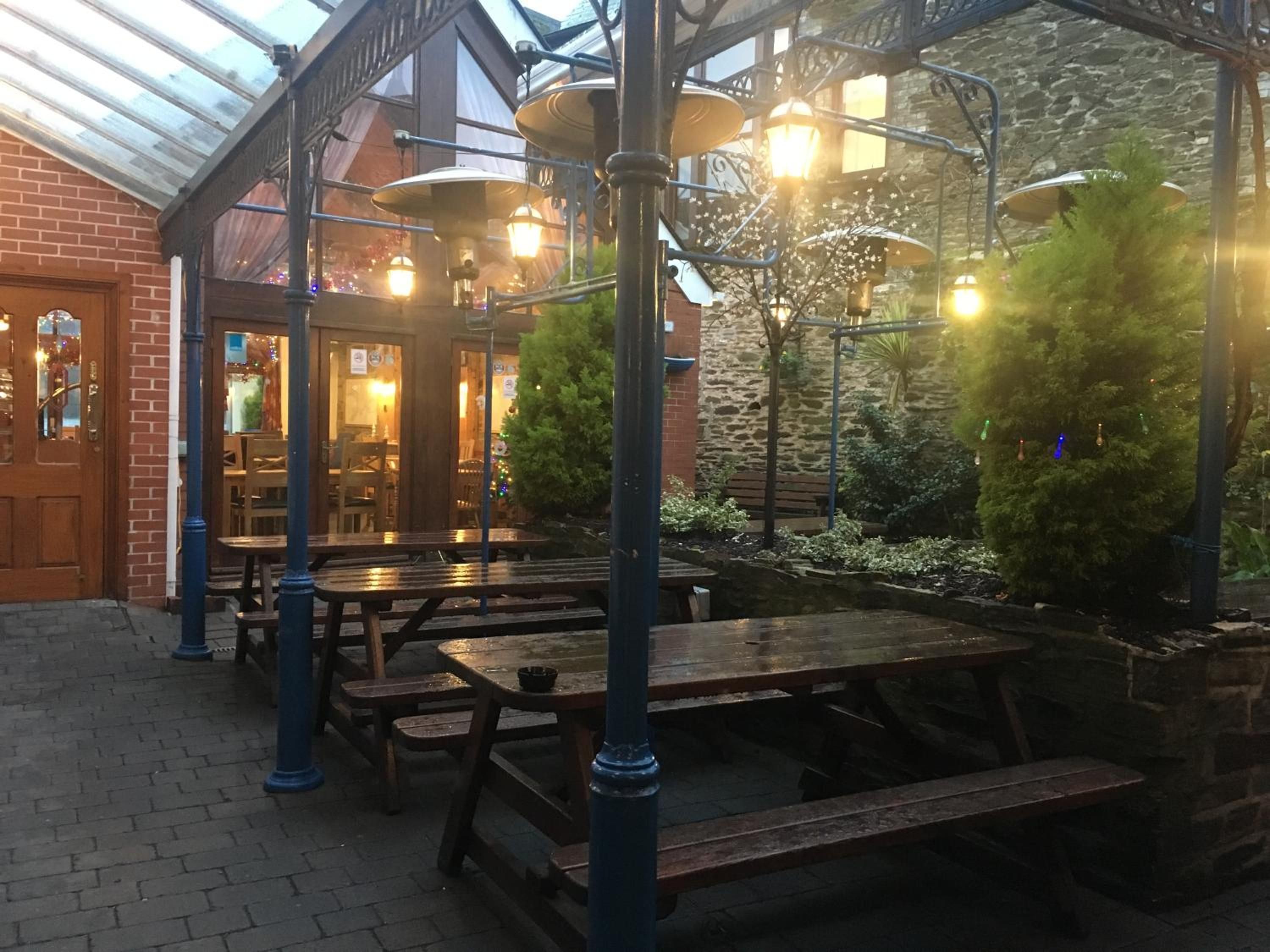An outdoor patio with wooden tables and benches, a brick wall, and a glass roof structure.