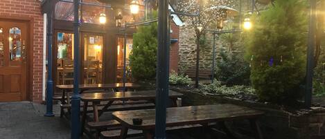An outdoor patio with wooden tables and benches, a brick wall, and a glass roof structure.