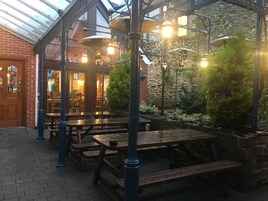 An outdoor patio with wooden tables and benches, a brick wall, and a glass roof structure.