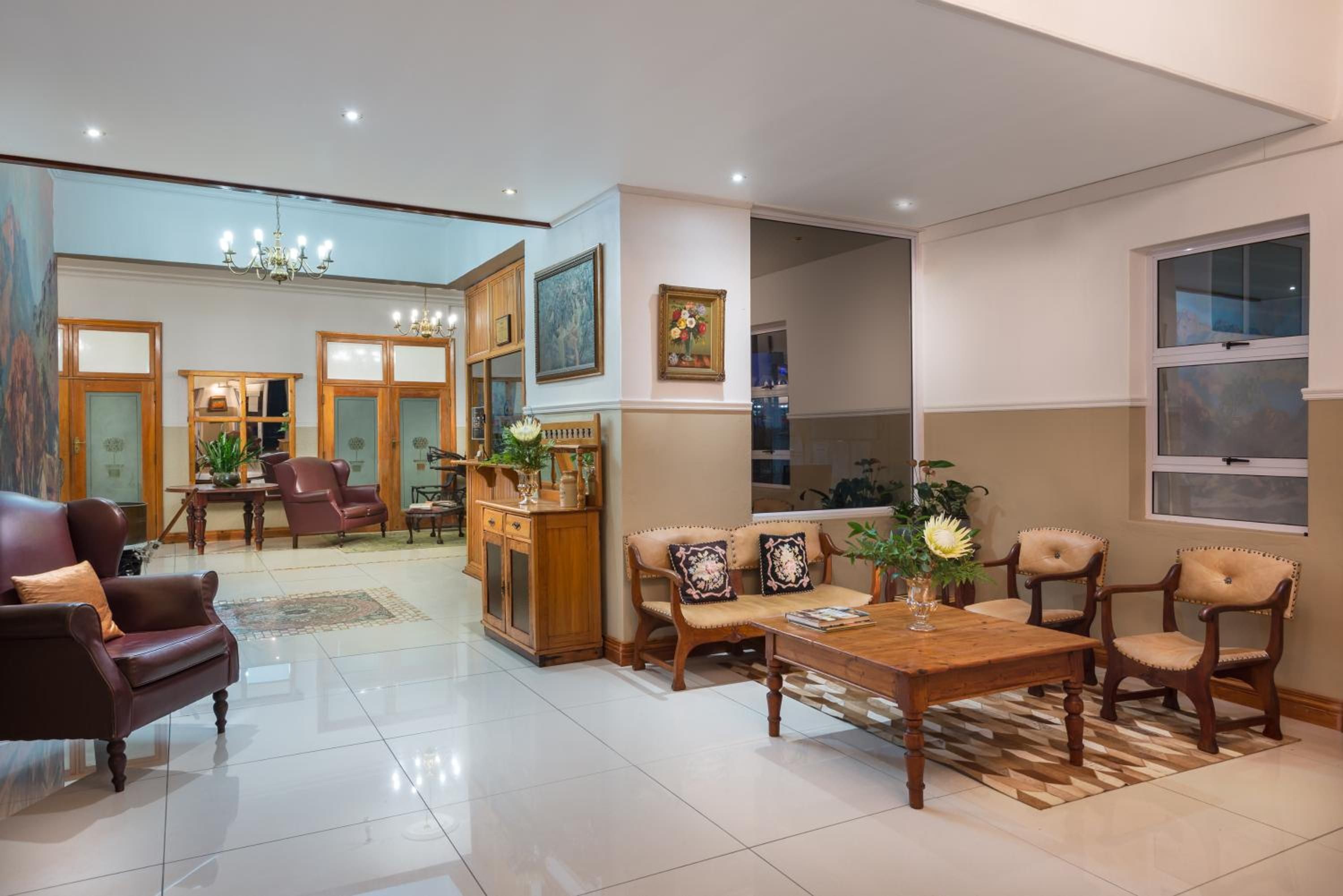 A spacious living room with a large wooden coffee table, leather armchairs, and a sofa. There is a dining area with a wooden cabinet and a chandelier.