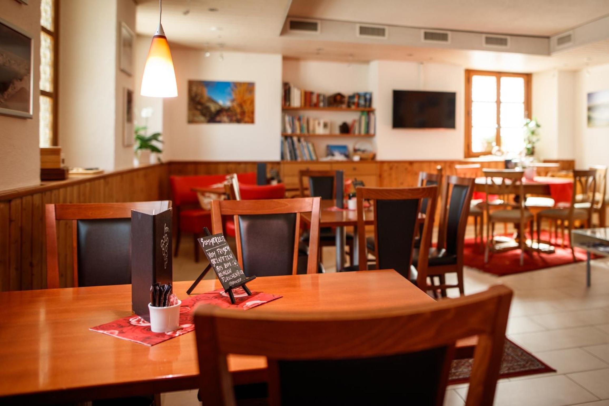 A restaurant interior with wooden tables and chairs, a blackboard menu, and a bookshelf in the background.