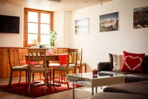 A cozy living room with a wooden dining table, chairs, a red rug, a glass coffee table, and a leather sofa with a heart-shaped pillow. - Hotel Nufenen (Obergoms)