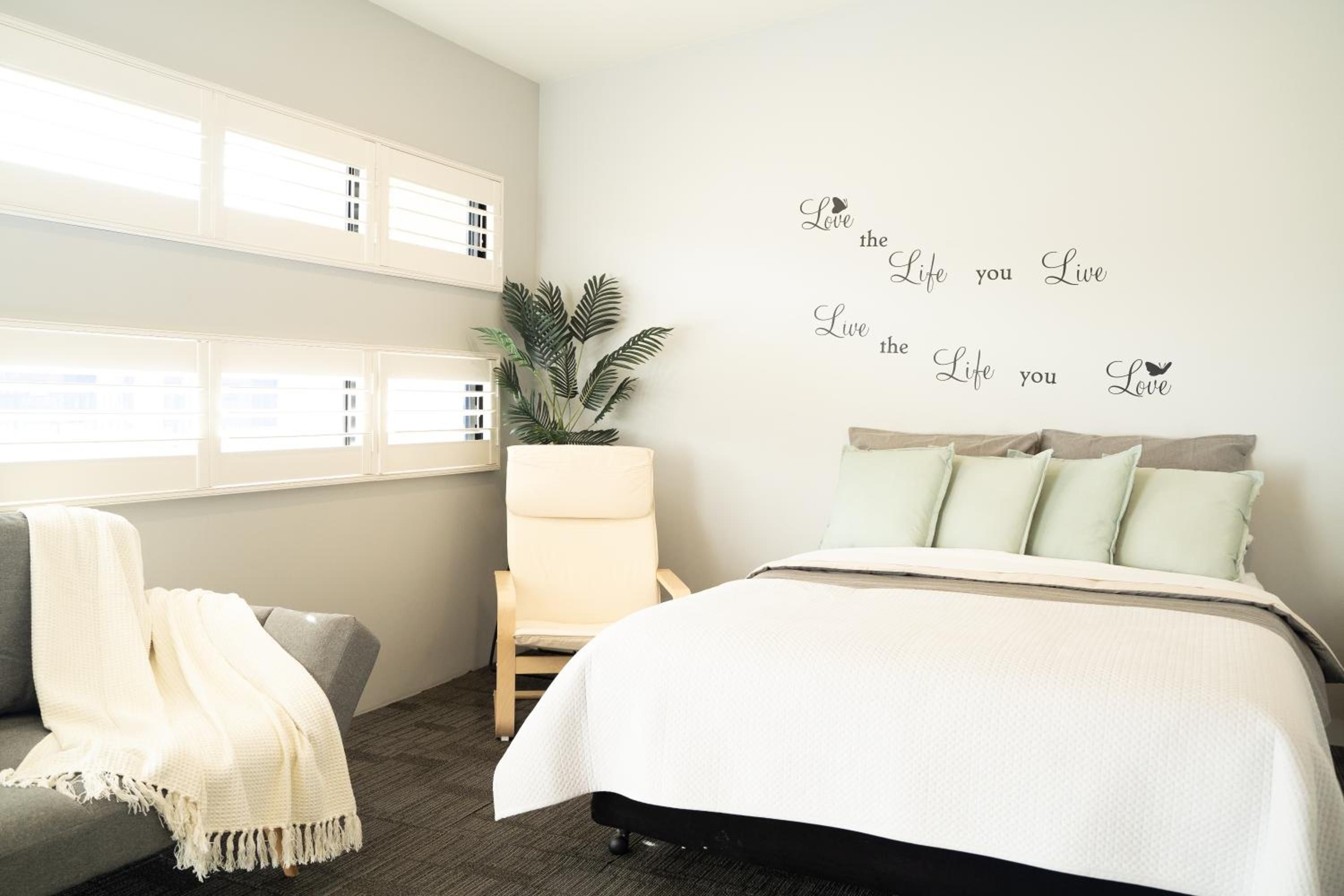 A bedroom with a bed, a chair, a potted plant, and a wall with a motivational quote.