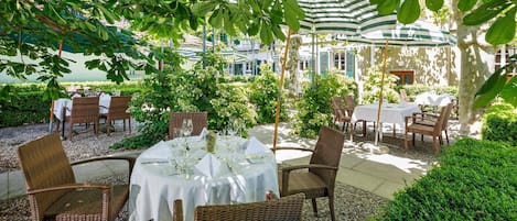 An outdoor dining area with tables and wicker chairs, surrounded by greenery and umbrellas.