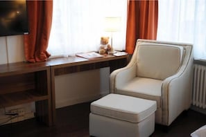 A hotel room with a wooden desk, a white armchair, a small ottoman, a television, and a teddy bear on the desk. - Falken Am Rotsee (Ebikon)