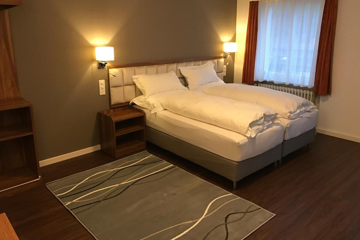 A hotel room with a large bed, bedside lamps, a nightstand, a wooden shelf, and a window with curtains.