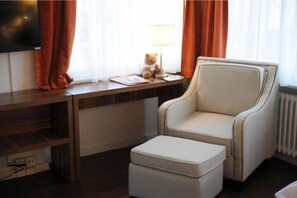 A hotel room with a wooden desk, a white armchair, a small ottoman, a television, and a teddy bear on the desk. - Falken Am Rotsee (Ebikon)