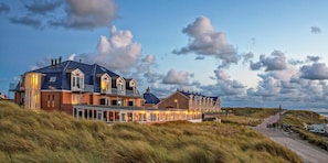 A large, multi-story building with a prominent roofline and multiple windows, situated on a grassy hill with a clear sky. - Strandhotel Noordzee (De Koog)