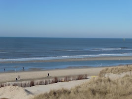 Ein Strand mit blauem Himmel, einem Leuchtturm in der Ferne und Menschen, die am Ufer entlanggehen.