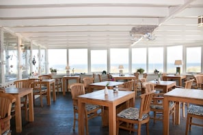 A spacious dining area with wooden tables and chairs, large windows offering a view of the sea, and a ceiling with a decorative bird sculpture. - Strandhotel Noordzee (De Koog)