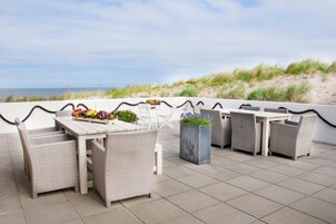 Una terraza en la azotea con mobiliario de mimbre, una mesa con un arreglo floral y vistas a la playa y las dunas.