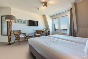 Una habitación de hotel con una cama grande, dos sillas de mimbre, una mesita, un televisor de pantalla plana y un ventanal con cortinas.