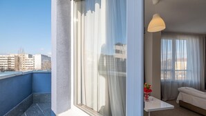 Ein Balkon mit Blick auf Gebäude, ein Tisch in Weiß mit einer Vase und ein Bett mit weißer Bettdecke.