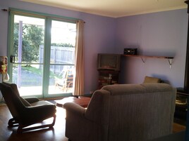 Una sala de estar con puerta corrediza de vidrio que da a un jardín, un televisor sobre un mueble bajo y un sillón mecededor.