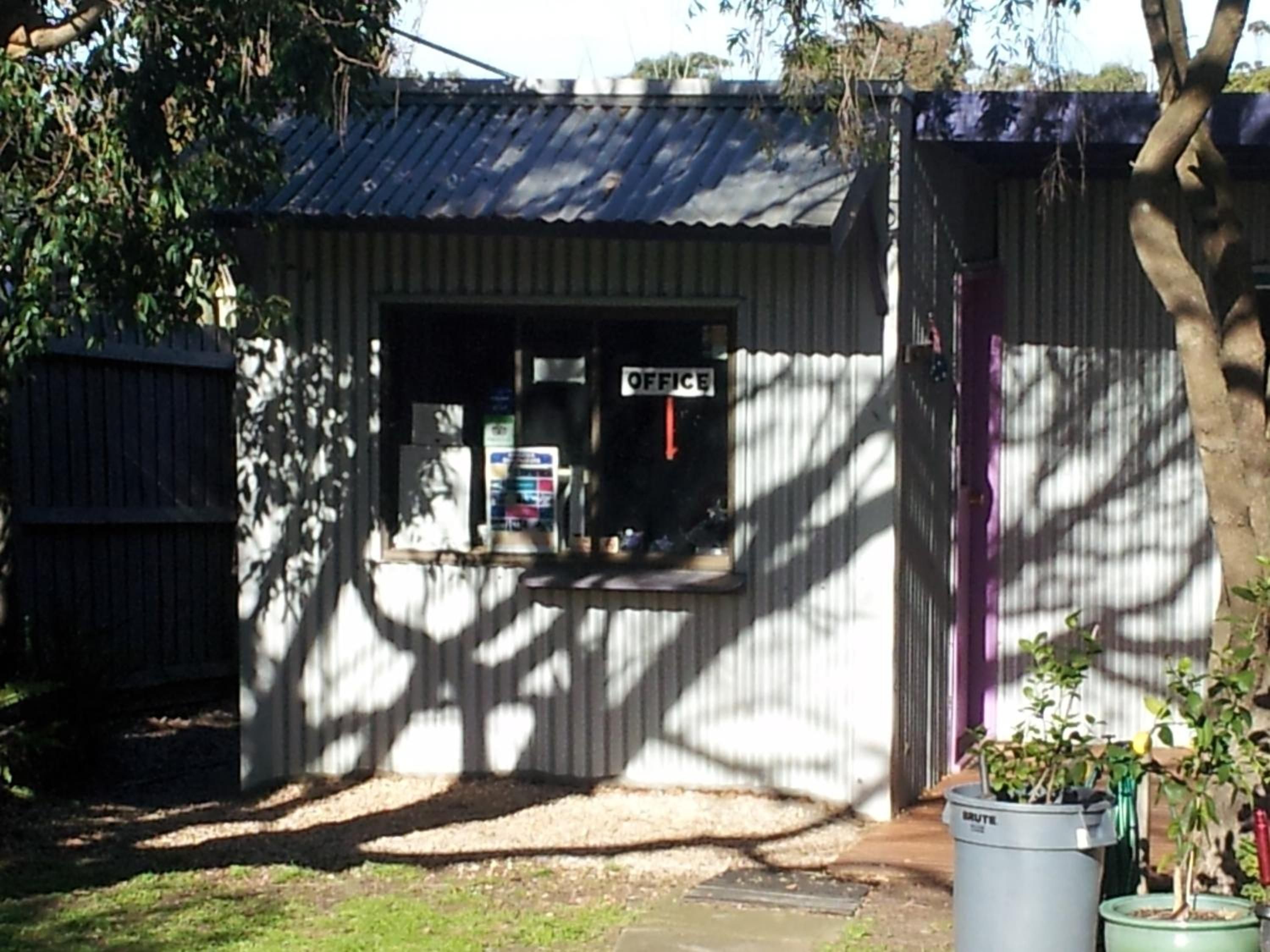 A small office with a corrugated metal roof, a window with an 'OFFICE' sign, and a potted plant in the foreground.