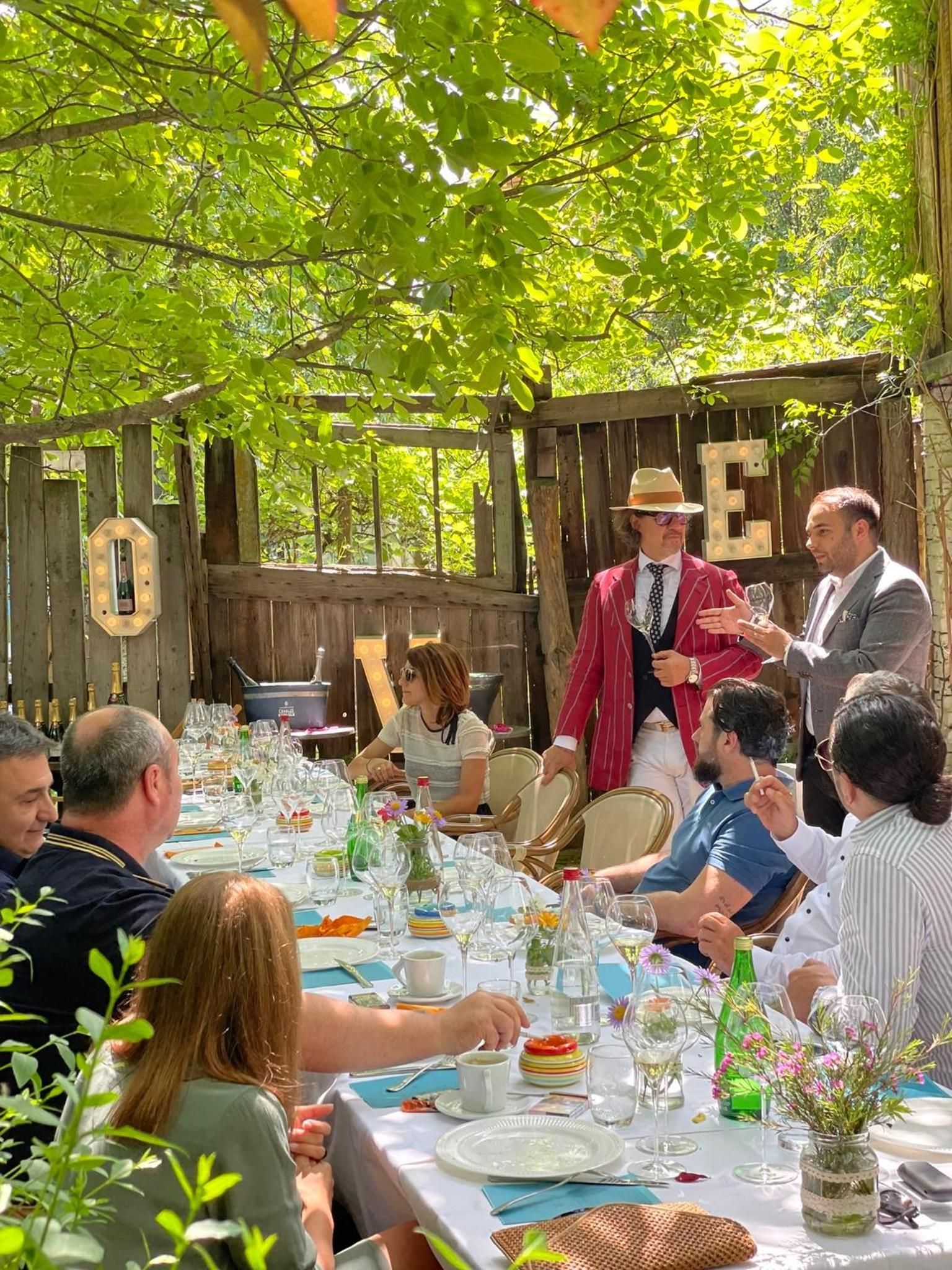 Un área de comedor al aire libre con mesas dispuestas para una comida, rodeada por una valla de madera y follaje.