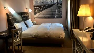 A hotel room with a bed, bedside table, lamp, chair, desk, and a cityscape painting on the wall.