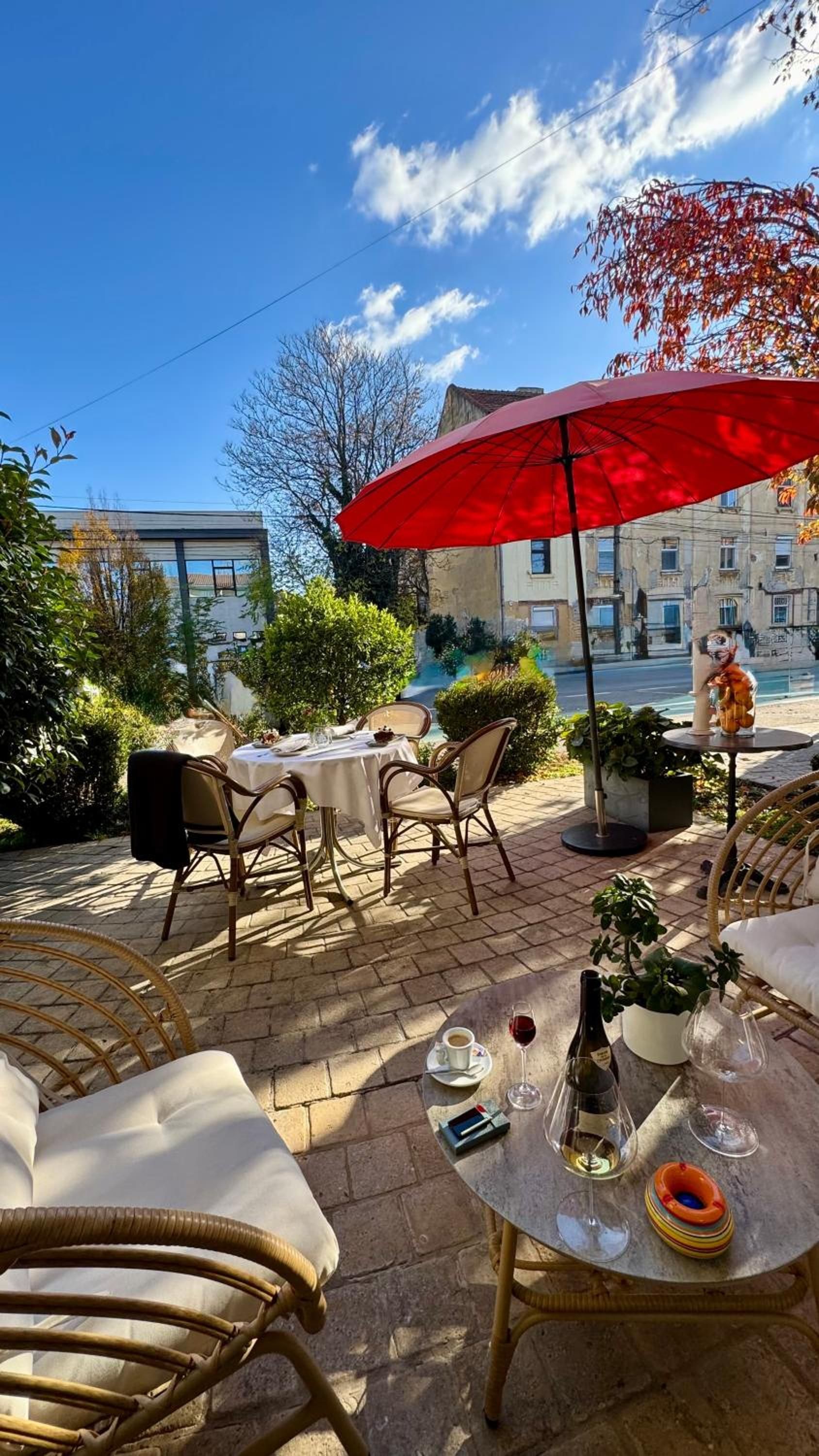 An outdoor café with tables and chairs, a red umbrella, and a view of a street with buildings.