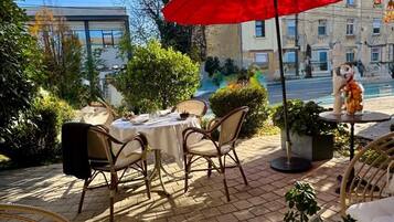 An outdoor café with tables and chairs, a red umbrella, and a view of a street with buildings.