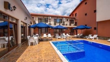 A pool area with lounge chairs and umbrellas, a tiled pool, and a building with multiple windows and balconies.