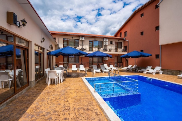 Un área de piscina con sillones de descanso y sombrillas, una piscina con azulejos y un edificio con múltiples ventanas y balcones.