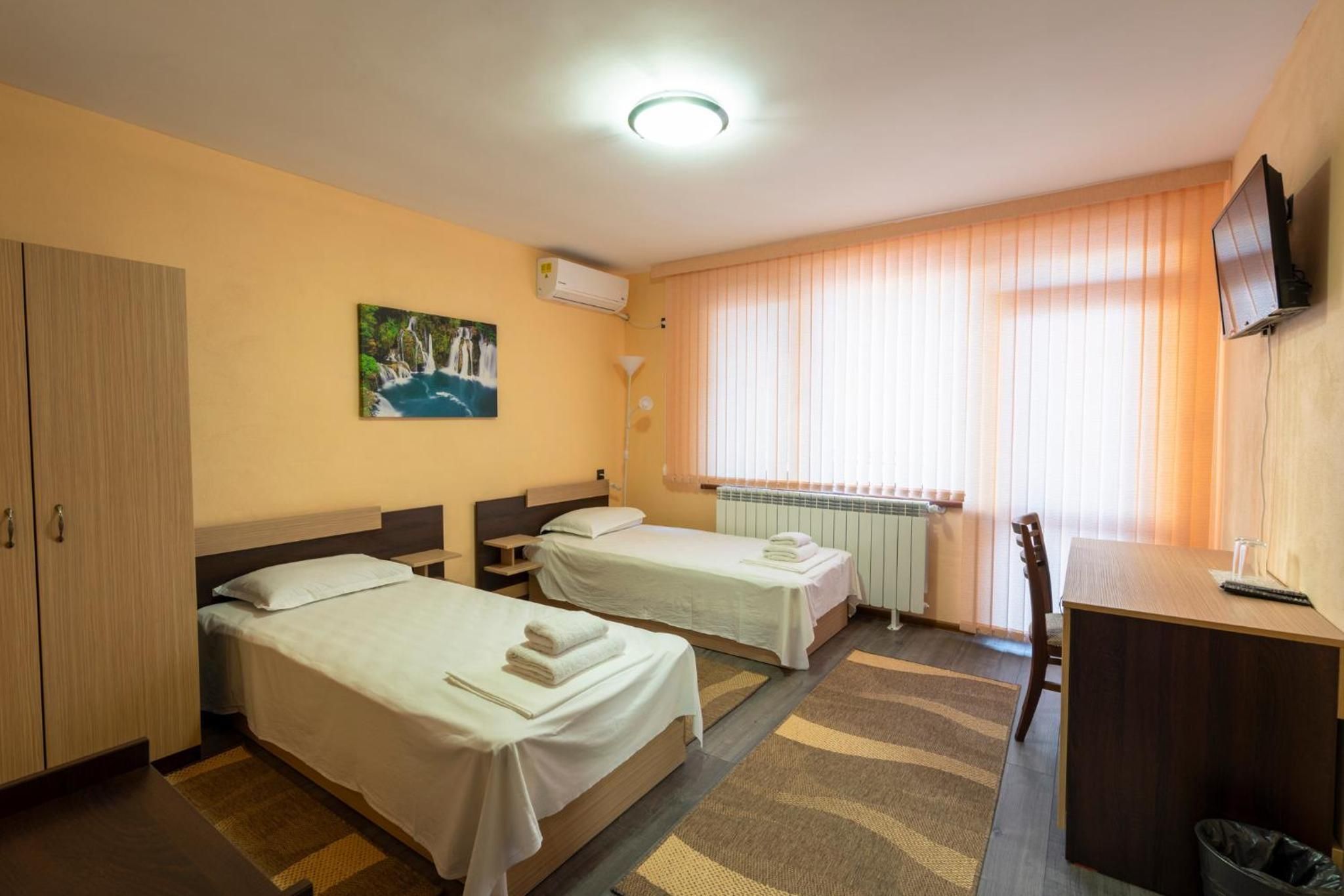 A hotel room with two beds, a desk, a chair, a TV, a radiator, and a painting on the wall.