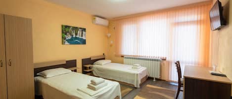 A hotel room with two beds, a desk, a chair, a TV, a radiator, and a painting on the wall.
