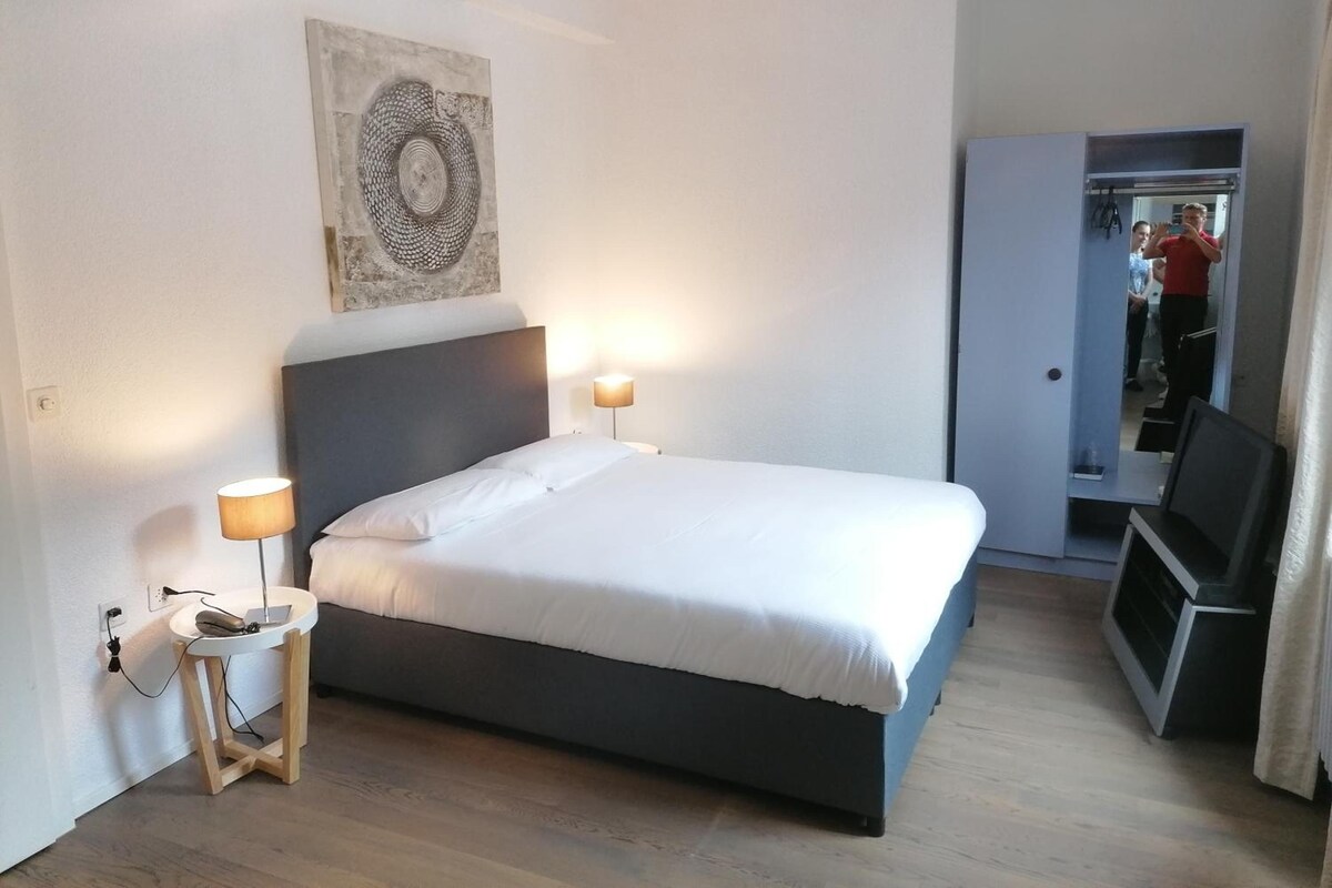 Ein Schlafzimmer mit einem Bett, Nachttischlampen, einem kleinen Tisch, einem Wandbild und einer Tür zu einem Badezimmer.