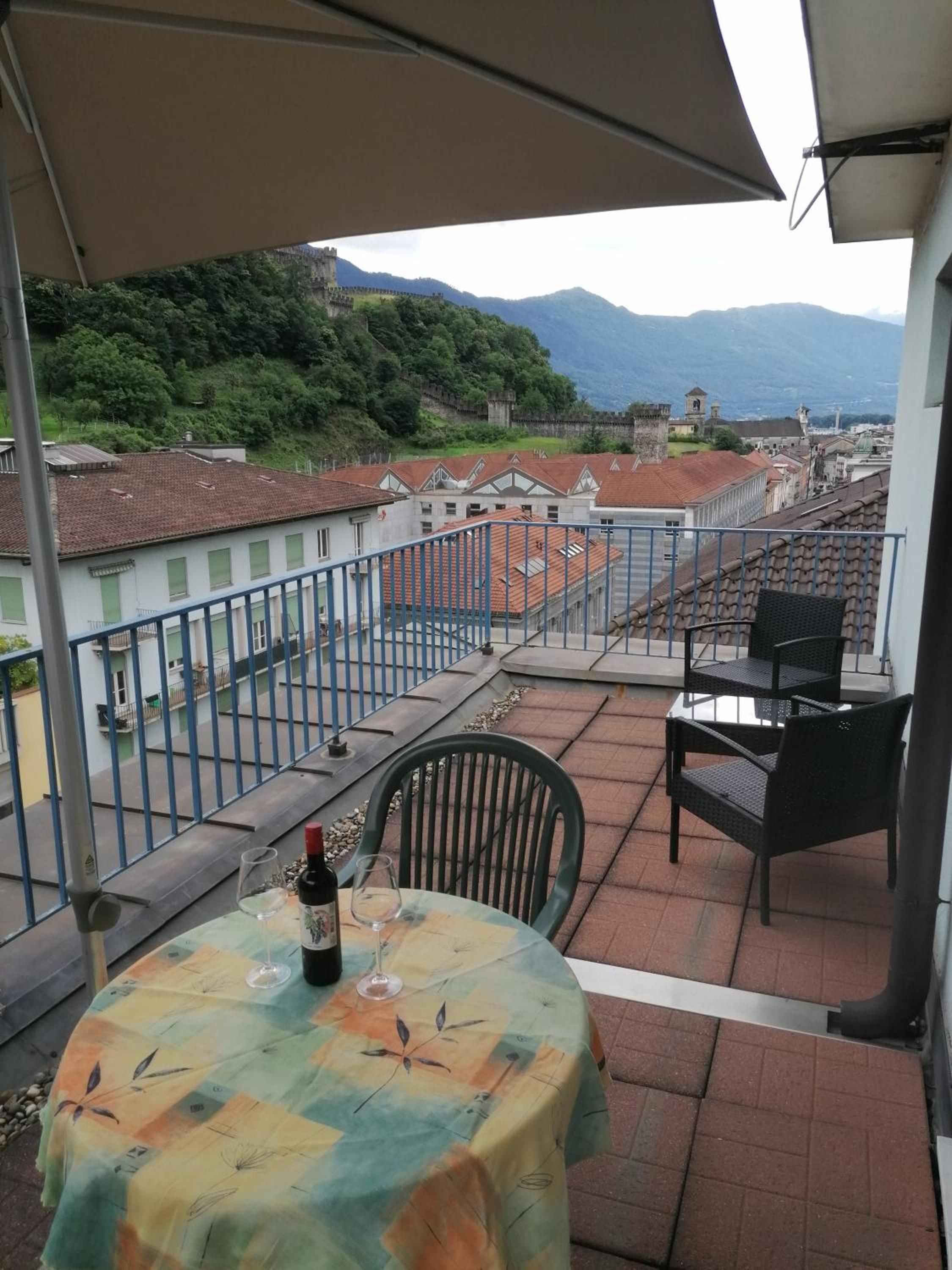 Eine Terrasse mit Tisch, Stühlen und einer Weinflasche. Von der Terrasse hat man einen Blick auf eine Stadtlandschaft mit Gebäuden und einer Hügelkette in der Ferne.