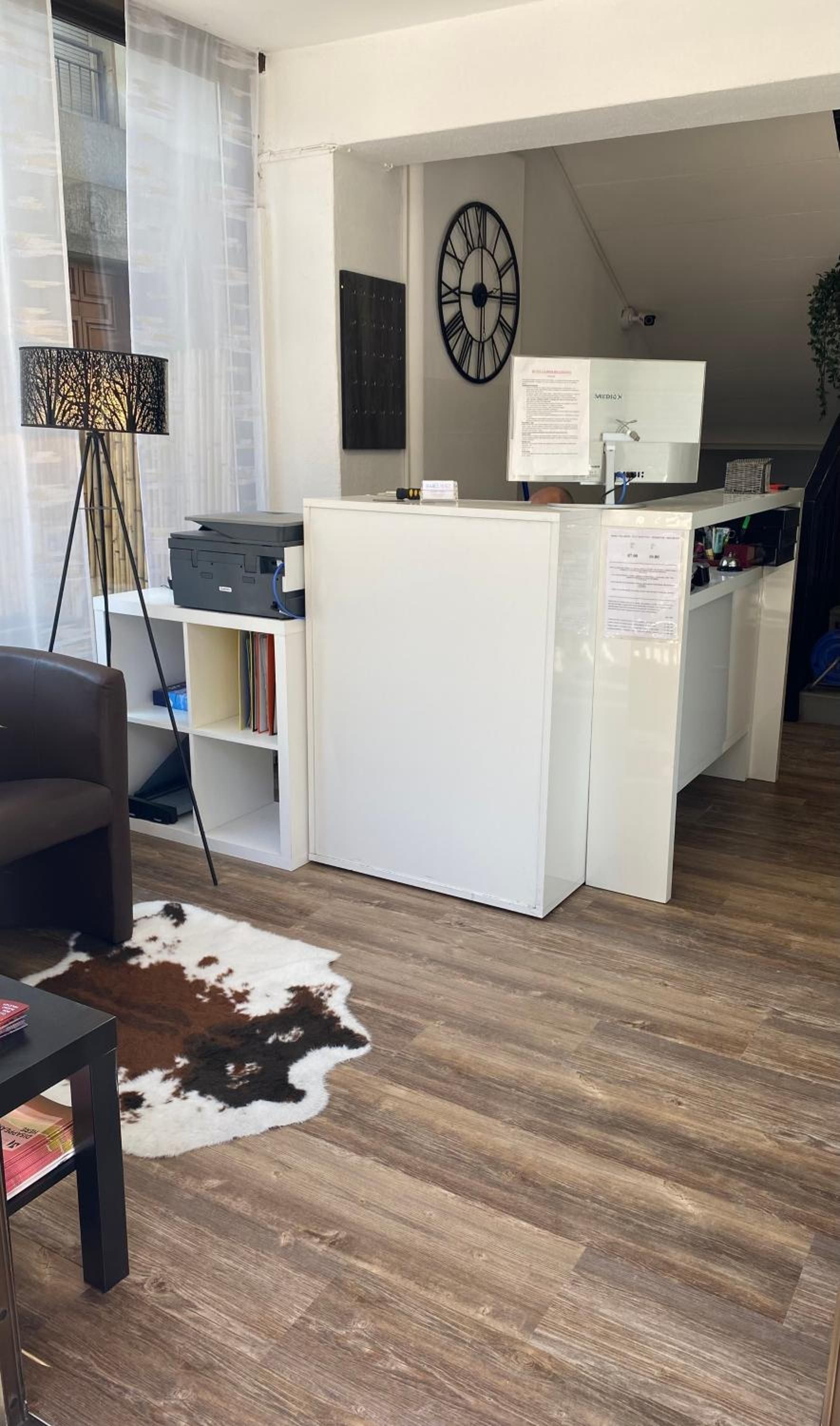 Ein moderner Arbeitsraum mit Schreibtisch, Computer und Drucker. An der Wand hängt eine Uhr und in der Ecke steht eine Pflanze.
