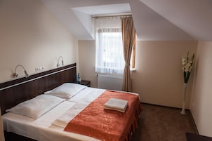 Una habitación de hotel con cama, mesita de noche, ventana con cortinas, radiador y un jarrón con flores.